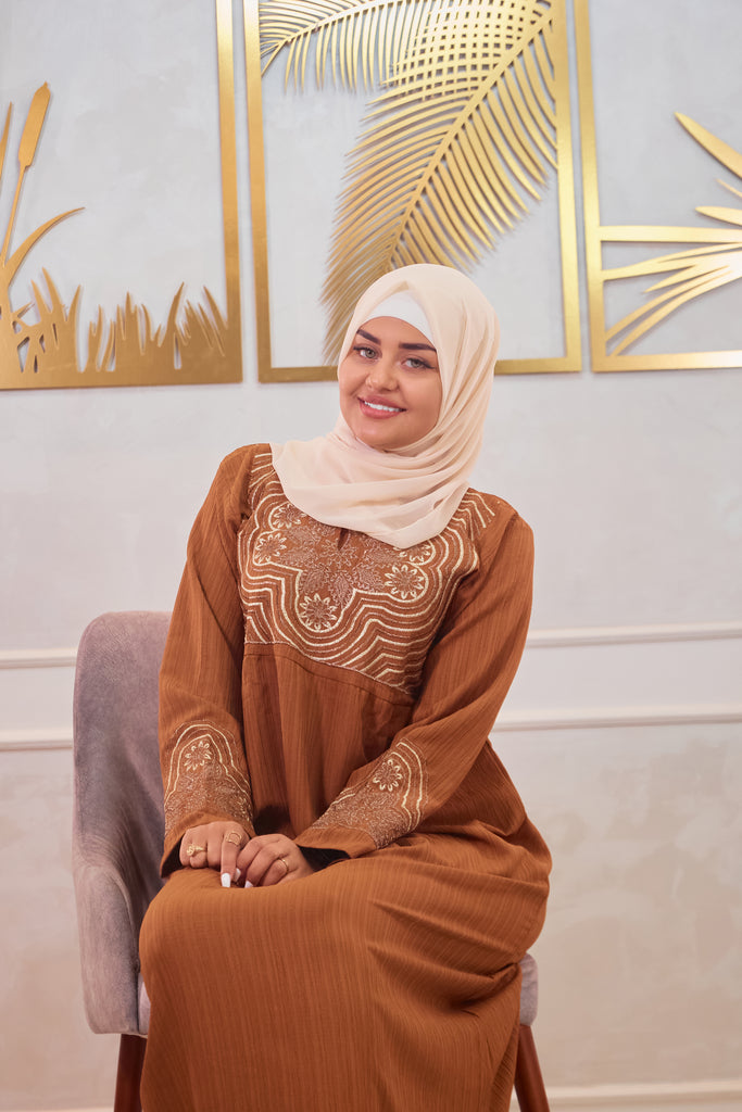 Woman in a brown luxury abaya dress with intricate patterns sitting on a chair against a decorative wall waring and cream cotton hijab.